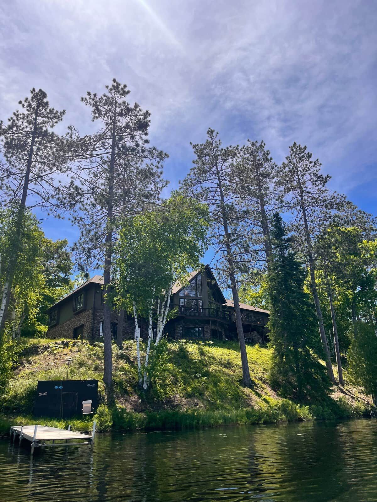 En stuga med en grund av sten, stora fönster och en balkong med utsikt över en sjö eller ett annat vattendrag.