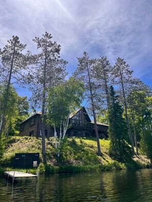 En stuga med en grund av sten, stora fönster och en balkong med utsikt över en sjö eller ett annat vattendrag.