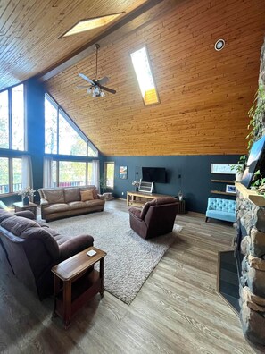 A spacious living room with a high ceiling, wooden beams, a large window, and a stone fireplace.