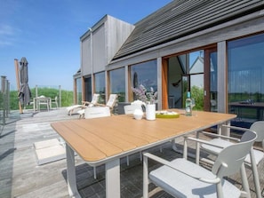 Eine moderne Außenterrasse mit Holztisch, weißen Stühlen, großen Fenstern und Blick auf die umliegende Landschaft.