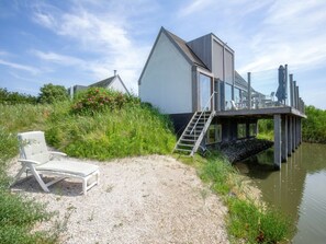 Ein modernes Haus mit Steg, der sich über das Wasser erstreckt, einem Kiesbereich mit Liegestuhl und einer Treppe zum Steg.