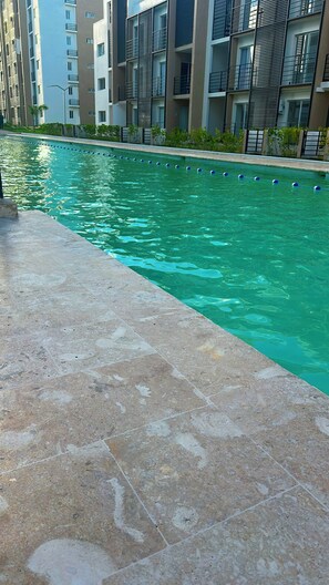 Ein Swimmingpool mit klarem Wasser, umgeben von einer gefliesten Liegefläche und modernen Gebäuden im Hintergrund.