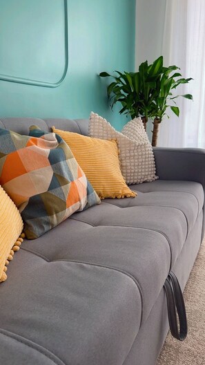 Ein graues Sofa mit vielen Kissen, einer Zimmerpflanze und einem Fenster mit Vorhängen.