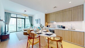 A modern kitchen with wooden cabinets, a dining area with a white table and chairs, and a large window offering a view of the city. - Lux 1 Bed Partial Sea View l Private Beach Access (Emaar Beachfront)