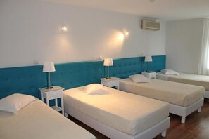 Une chambre d’hôtel avec trois lits simples, chacun doté d’un matelas blanc et d’oreillers assortis. On y trouve des tables de chevet avec des lampes, ainsi qu’un climatiseur fixé au mur.