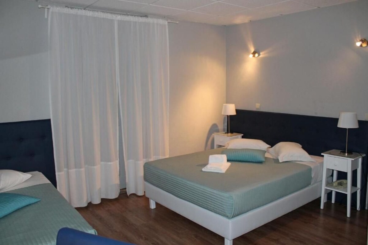 Une chambre d’hôtel avec un lit simple, deux tables de chevet, une petite table, une lampe et un rideau.