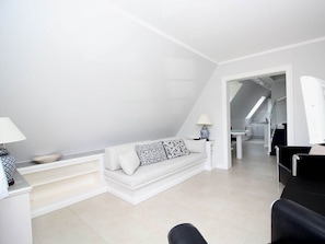 A modern living room with a white sofa, a small table with a lamp, and a black chair. A staircase leads to an upper level. - Island Love OG W3 (Sylt)