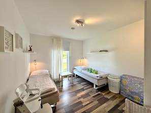 A bedroom with two beds, a window with curtains, a bedside table, a chair, and a wall-mounted shelf. - Holiday Apartment With 2 Bedrooms (Sierksdorf)