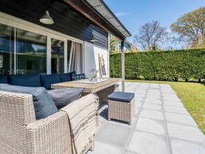An outdoor seating area with wicker furniture and cushions, a table with a placemat, and a view of a hedge and trees in the background. - Holiday Home in Renesse With Swimming Pool (Renesse)