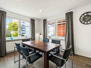 A modern dining area with a wooden table and metal chairs, large windows with curtains, and a wall clock. - Holiday Home With 2 Bedrooms (Krummhoern)