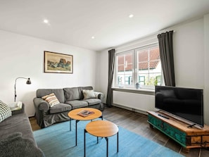 A modern living room with a grey sofa, two round wooden tables, a television on a wooden stand, and a window with curtains. - Holiday Home With 2 Bedrooms (Krummhoern)