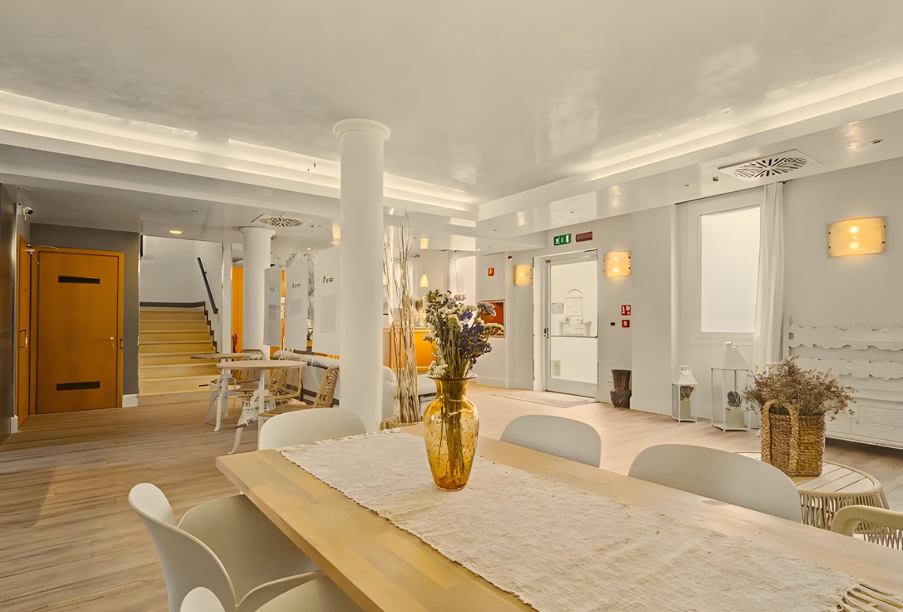 Un hall d’hôtel moderne avec un comptoir d’accueil en bois, des chaises blanches, un grand vase rempli de fleurs et un escalier menant à un autre espace.