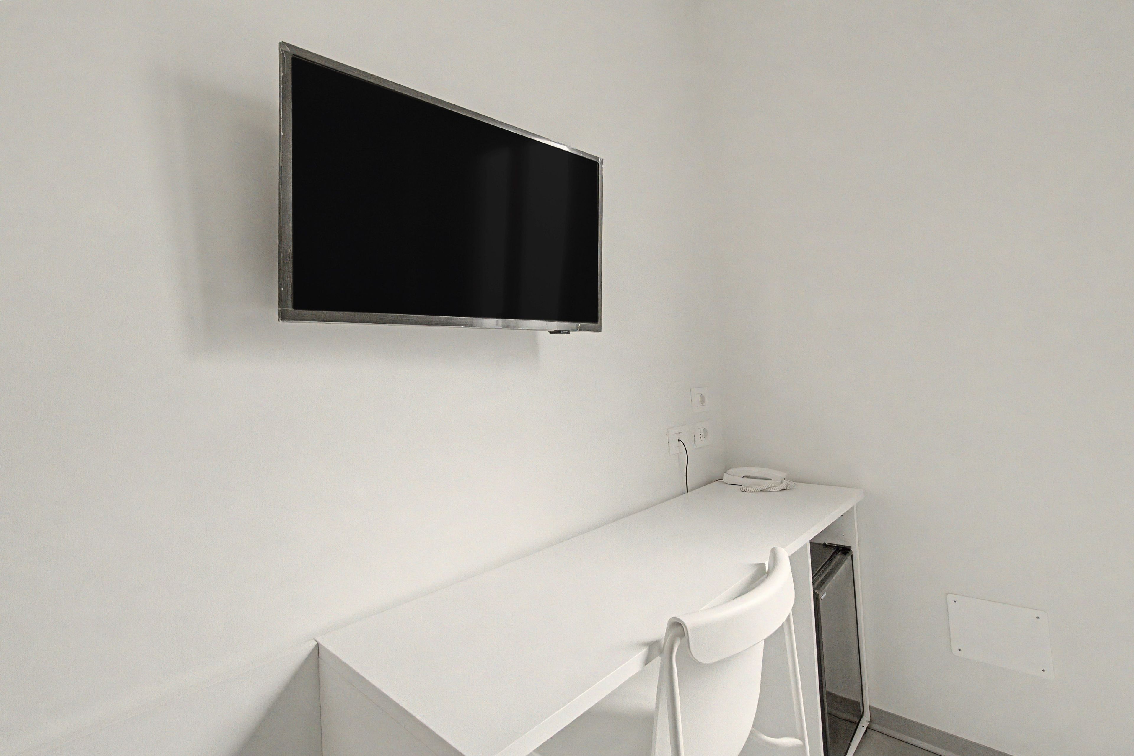 A white wall with a mounted television and a white counter with a telephone.