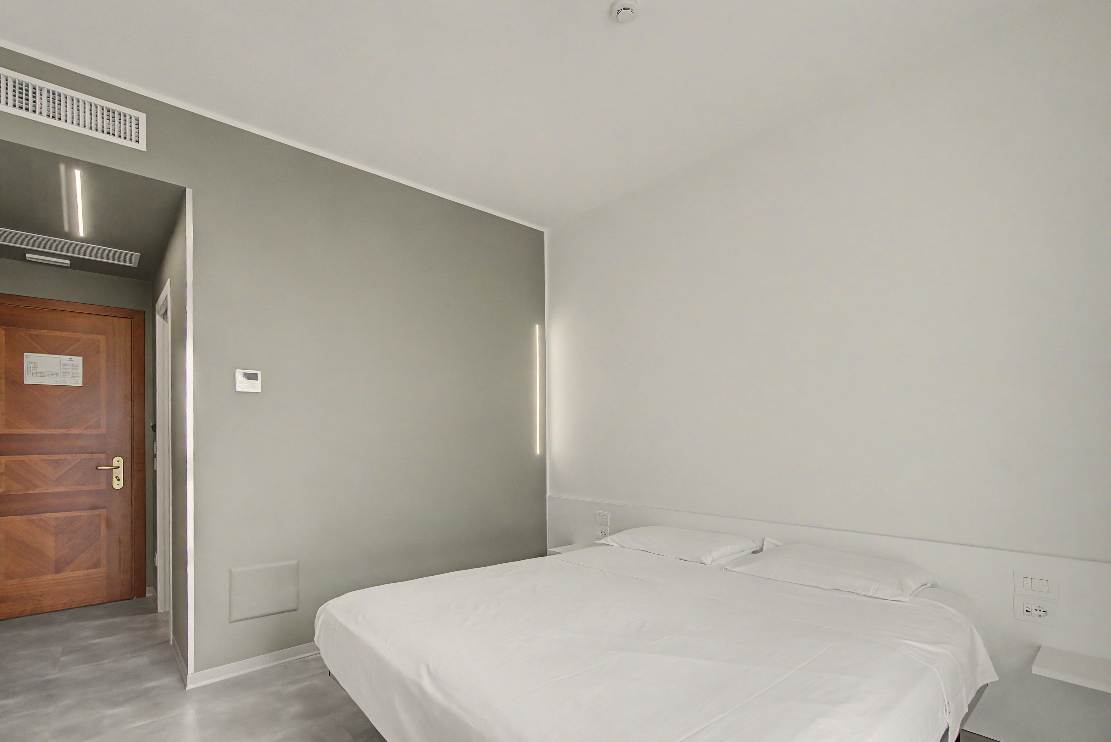 A hotel room with a bed, a door, a vent, and a light switch on the wall.