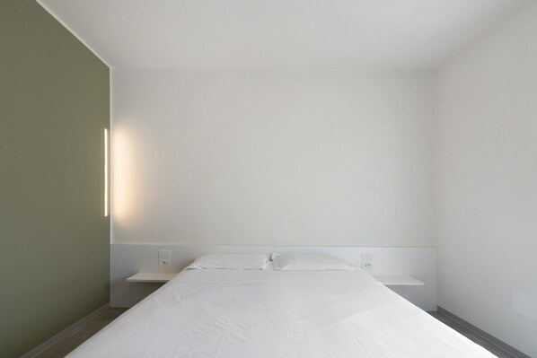 Une chambre d’hôtel minimaliste, avec un lit simple, des tables de chevet et un luminaire fixé au mur.
