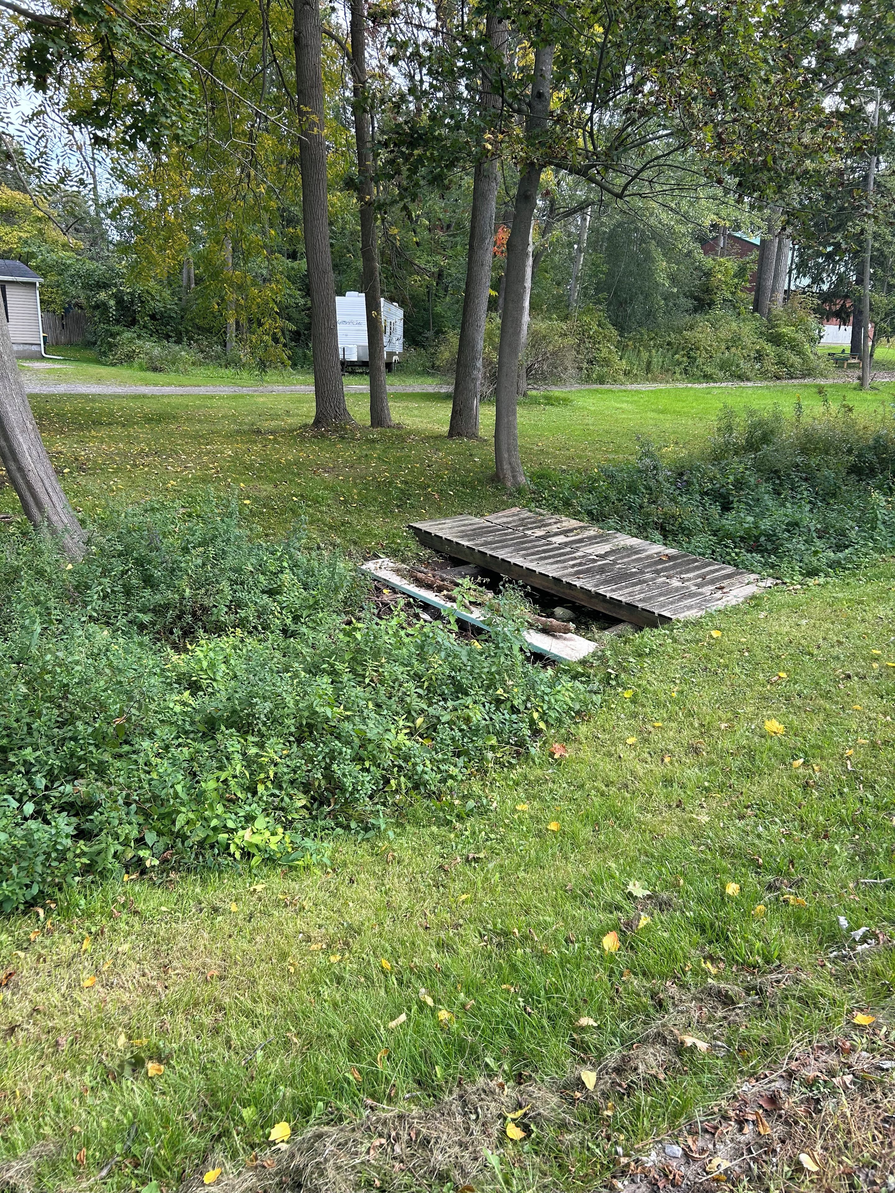 A grassy area with a wooden structure, surrounded by trees and a white trailer in the background.