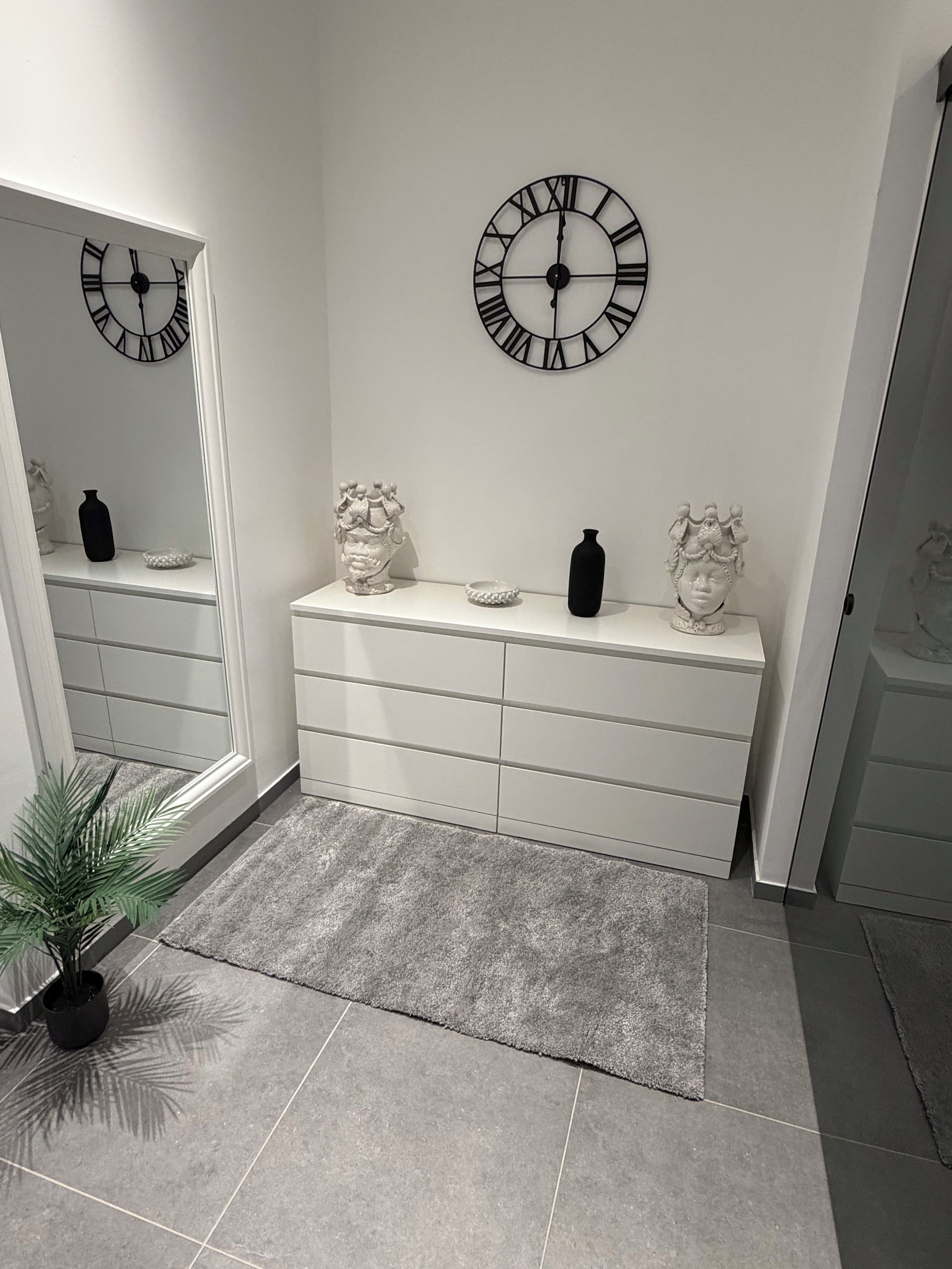 Un baño moderno con tocador blanco, dos relojes de pared, jarrones decorativos y una planta en maceta.