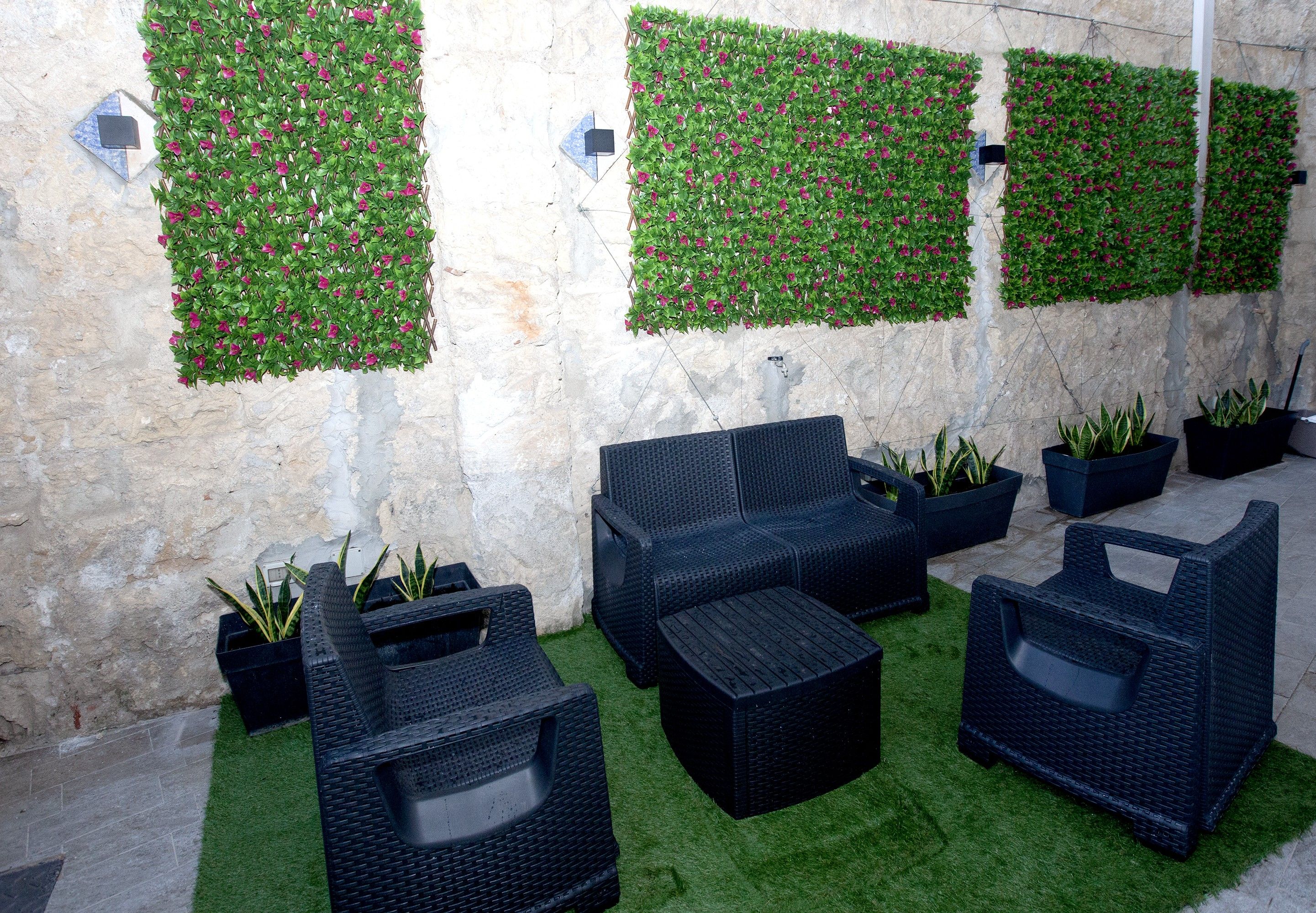 Un espace détente avec des meubles en osier noir, une pelouse artificielle verte et des plantes décoratives, le tout adossé à un mur en pierre.