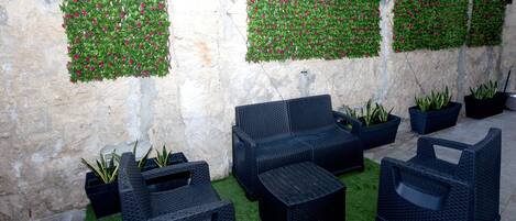 A seating area with black wicker furniture, green artificial grass, and decorative plants against a stone wall.