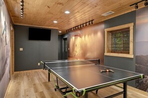 A ping pong table in a room with wooden flooring, a mounted television, and a window with blinds. - Secluded Yard • Pet-Friendly • Hot Tub (Sevierville)