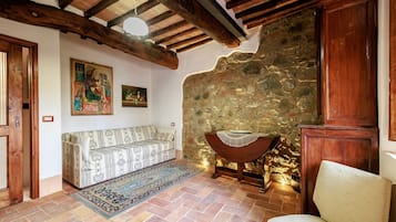 Ein gemütlicher Raum mit Steinwand, Holzbalkendecke, einem gemusterten Sofa, einem kleinen Tisch mit Spitzenbettdecke und einem Stuhl.
