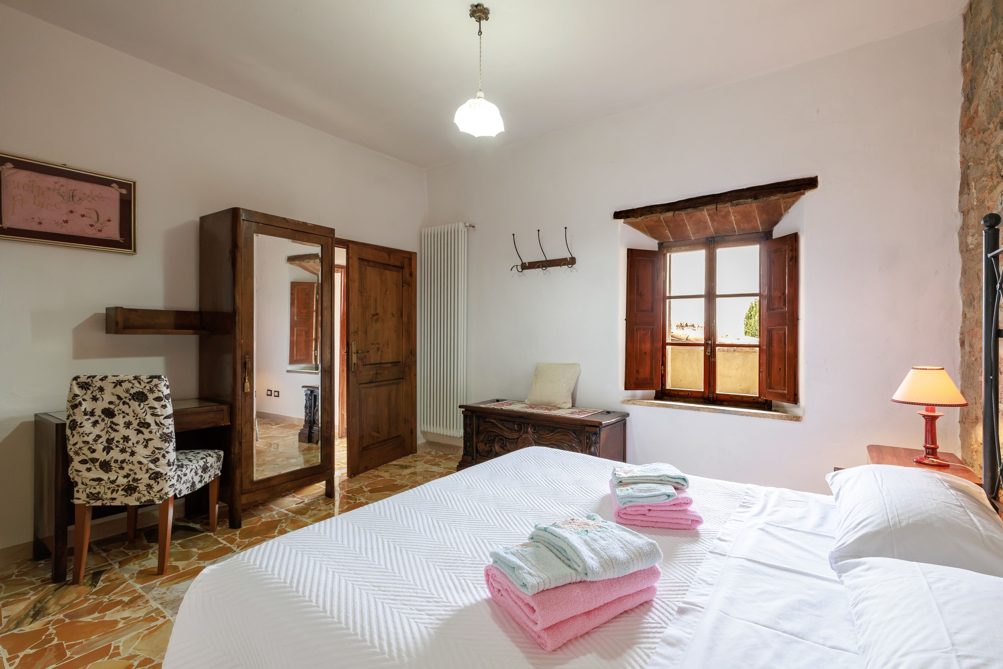 A bedroom with a bed, a chair, a desk, a radiator, a window with a view, and a hanging light fixture.