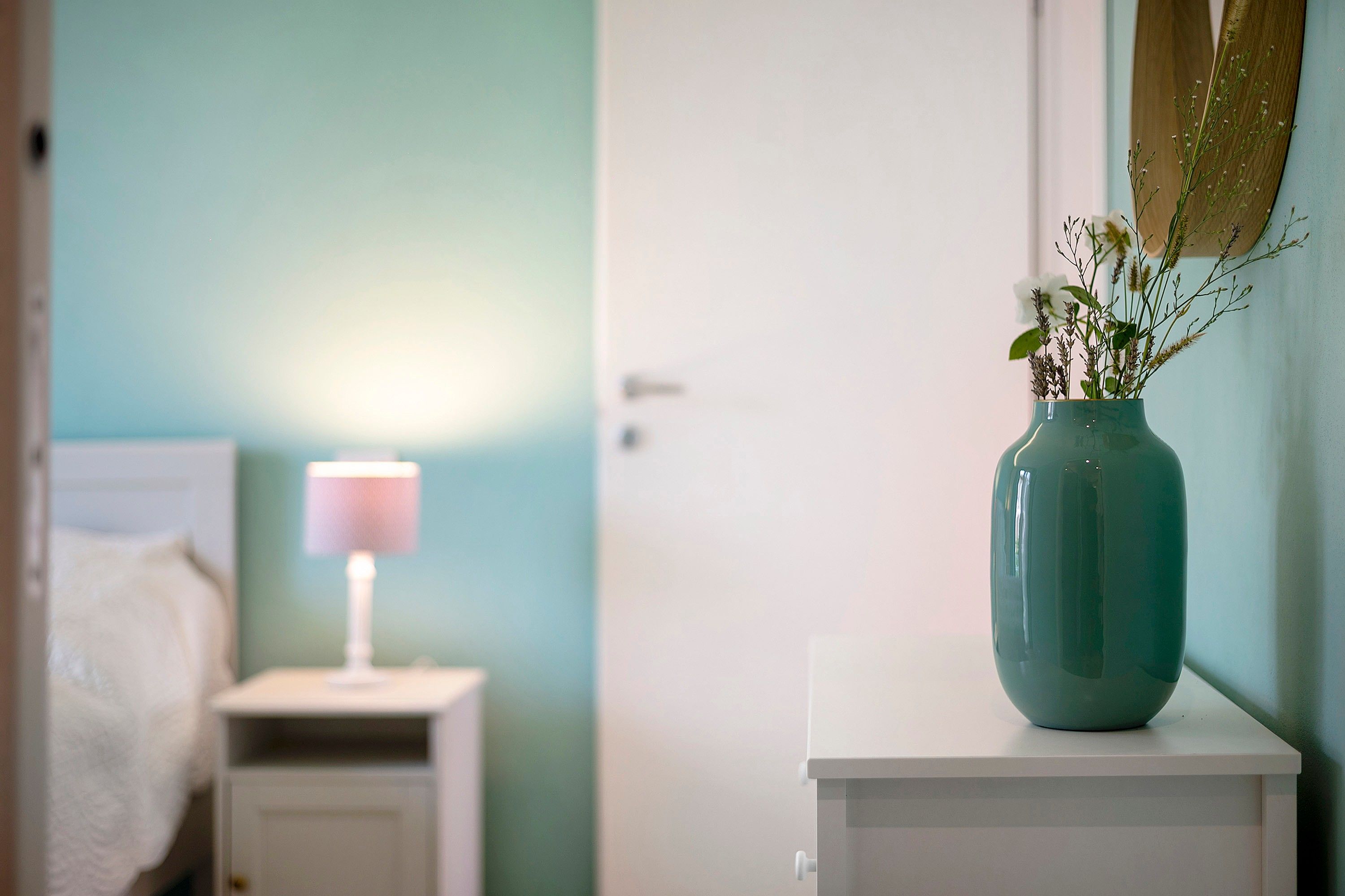 Una habitación con una mesita de noche, una lámpara y un florero con flores sobre un tocador.