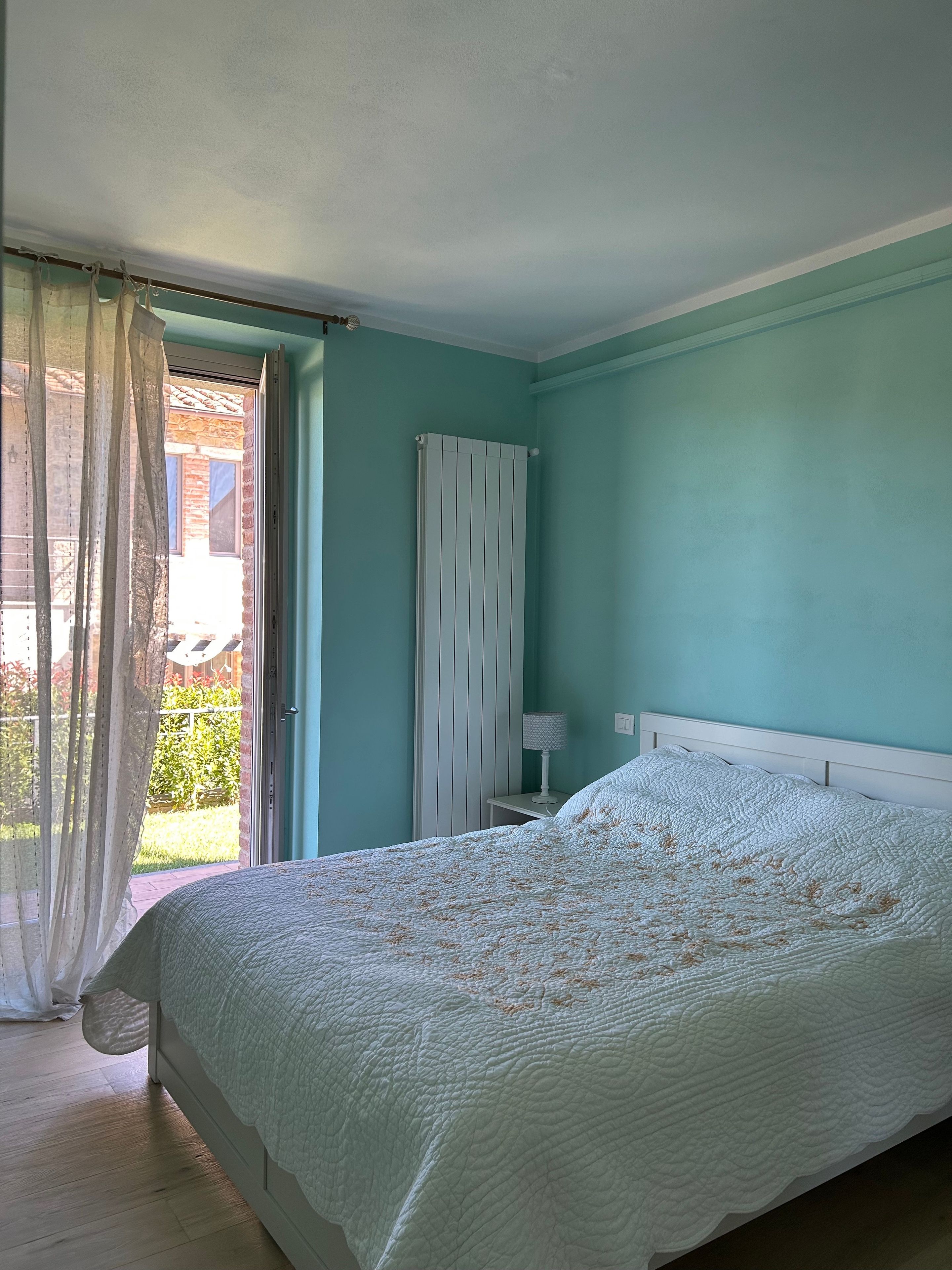 Ein Schlafzimmer mit einem Bett, einem Nachttisch, einem Heizkörper und Blick ins Freie.