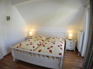 A bedroom with a white bed, two bedside tables with lamps, a radiator, and a wooden floor. - Holiday Home in Glowe With Swimming Pool (Glowe)