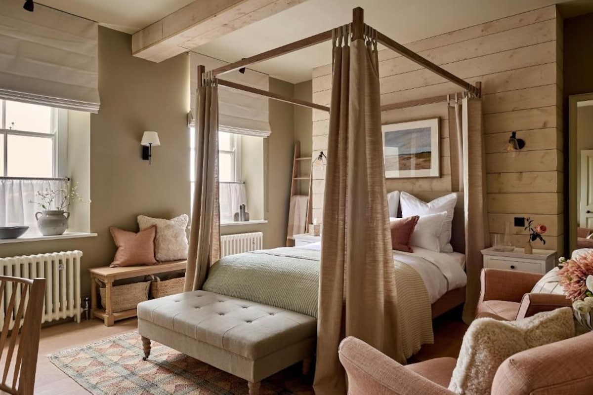 A bedroom with a four-poster bed, a wooden ceiling, a radiator, a bedside table with a lamp, and a window with curtains.