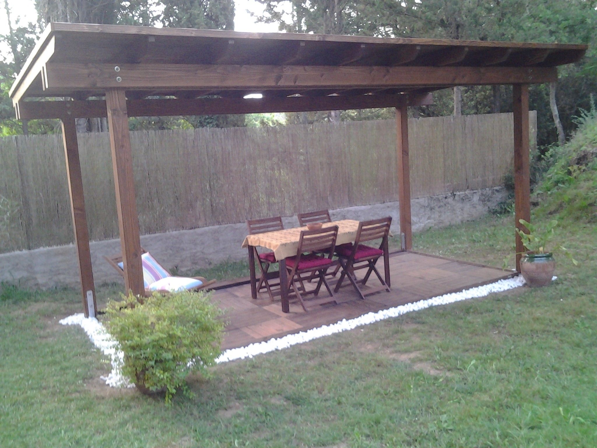 Un patio techado con una mesa y sillas de madera, rodeado por una cerca de madera y un jardín césped.