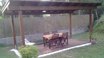 Un patio techado con una mesa y sillas de madera, rodeado por una cerca de madera y un jardín césped.