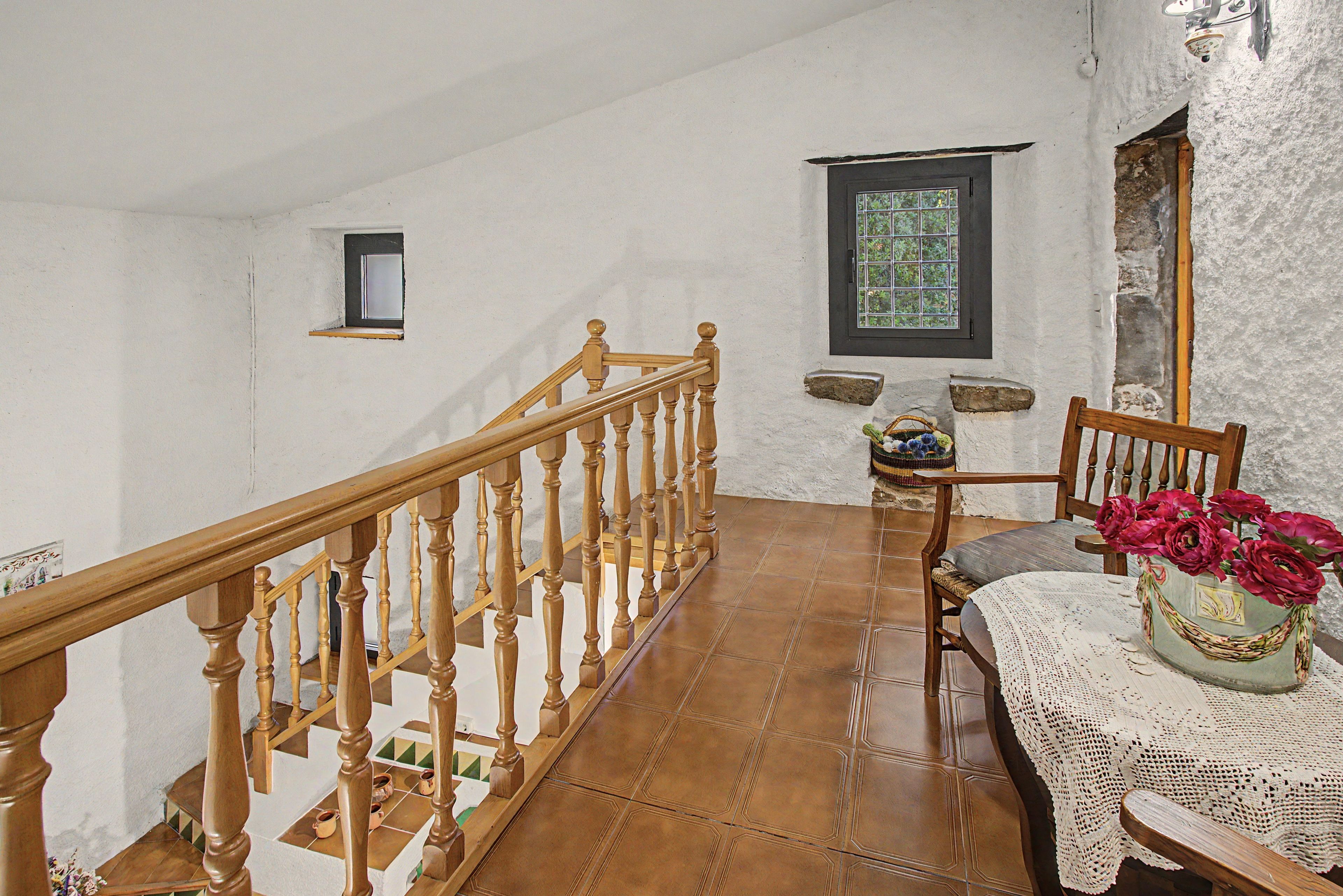 Una escalera con balaustres de madera que conduce a un rellano con una mesa y una silla, un ventanal con vistas, y un estante pequeño con objetos decorativos.