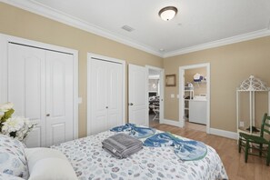 Un dormitorio con cama, dos puertas, cómoda, silla y una mesita de noche con lámpara.