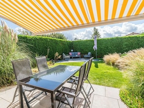 An outdoor patio with a glass table and chairs, a striped awning overhead, and a well-maintained hedge in the background. - Holiday Home in Renesse With Swimming Pool (Renesse)