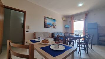 A dining area with a table set for two, a painting on the wall, and a view through a window.