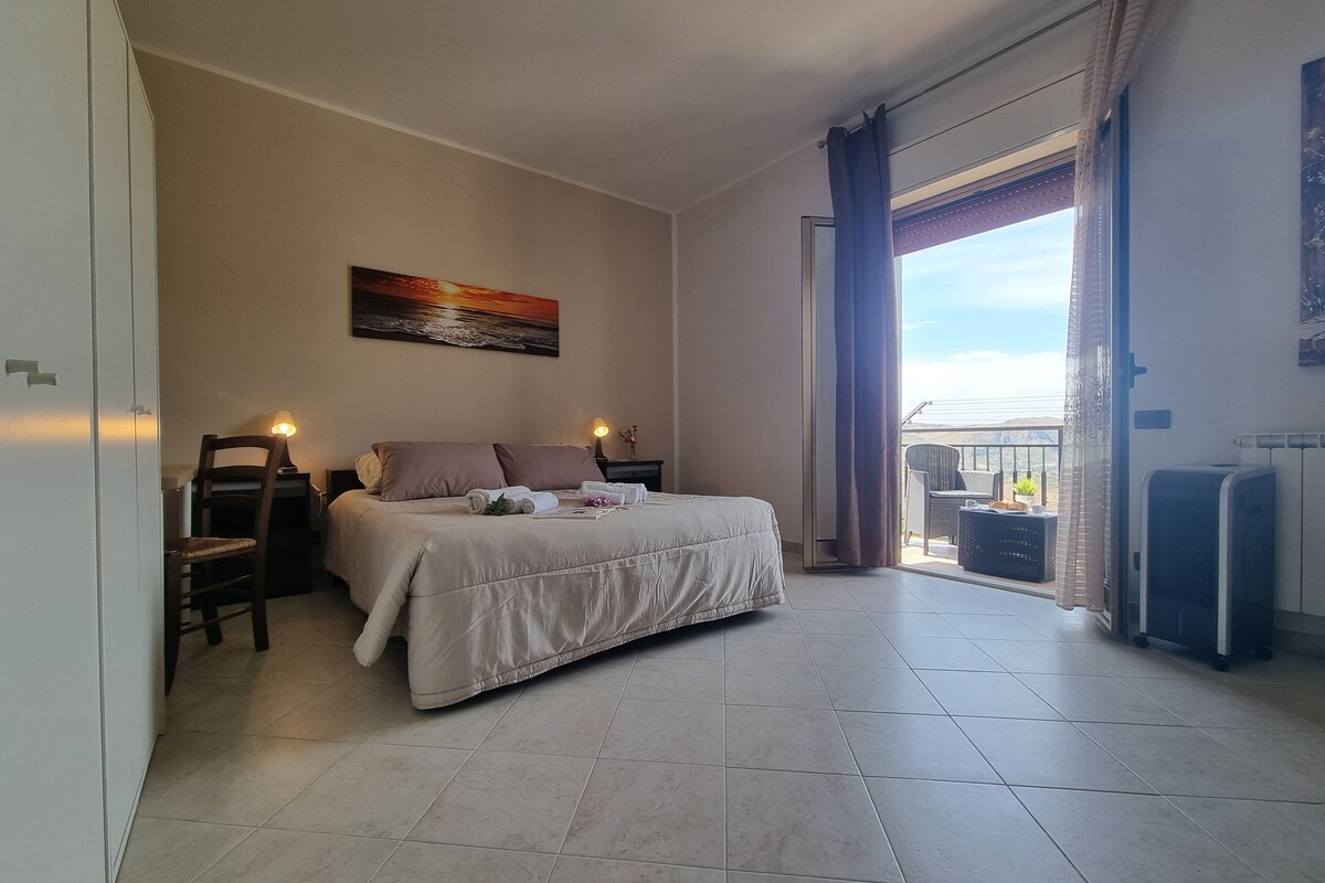 Ein Schlafzimmer mit Bett, Sessel, Schreibtisch, Lampe, einem Gemälde und Blick ins Freie.