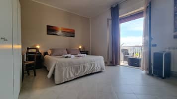 Ein Schlafzimmer mit Bett, Sessel, Schreibtisch, Lampe, einem Gemälde und Blick ins Freie.