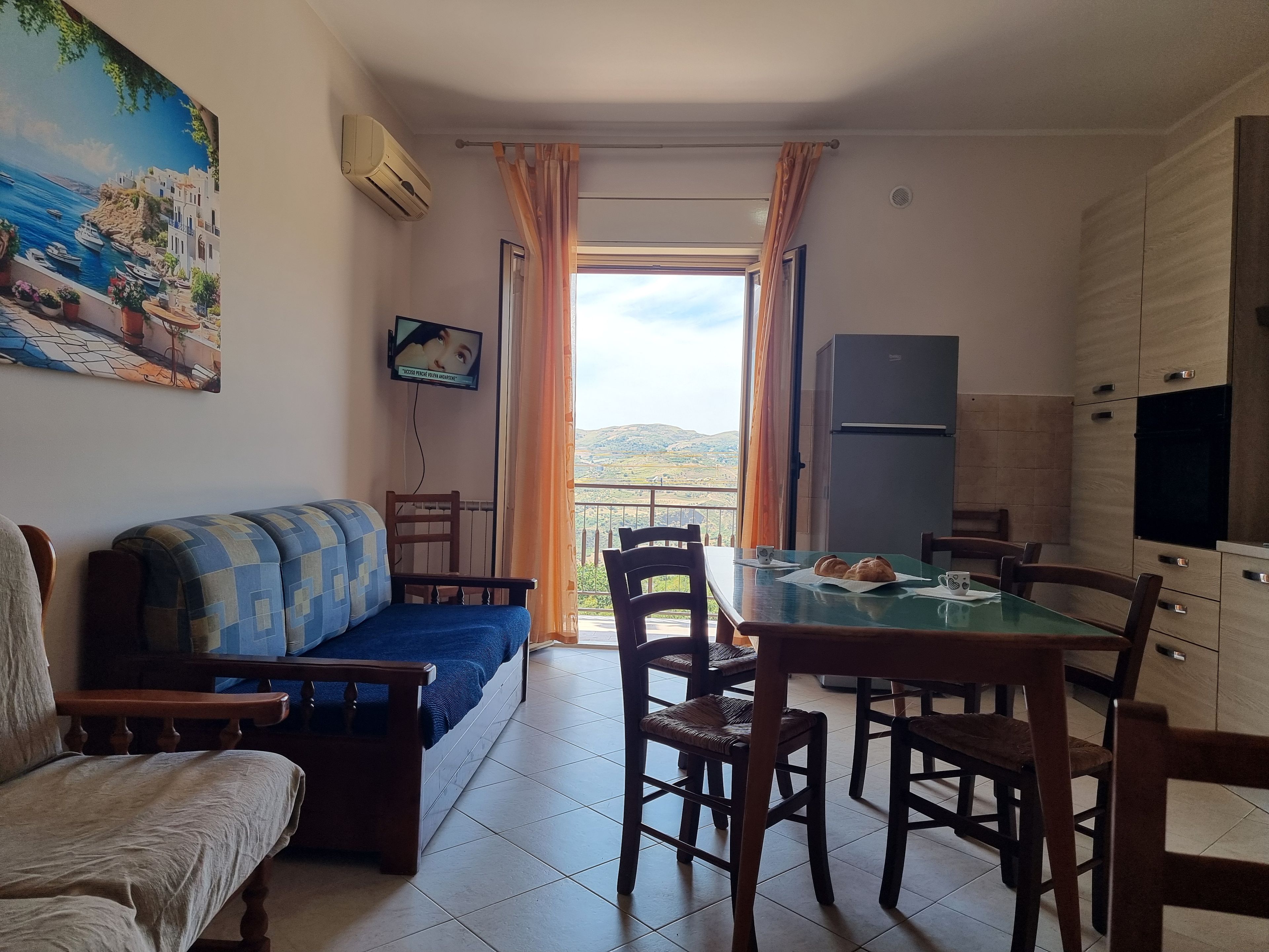 Una sala de estar con sofá, una mesa de comedor con sillas, un televisor montado en la pared y vistas al exterior a través de un ventanal.