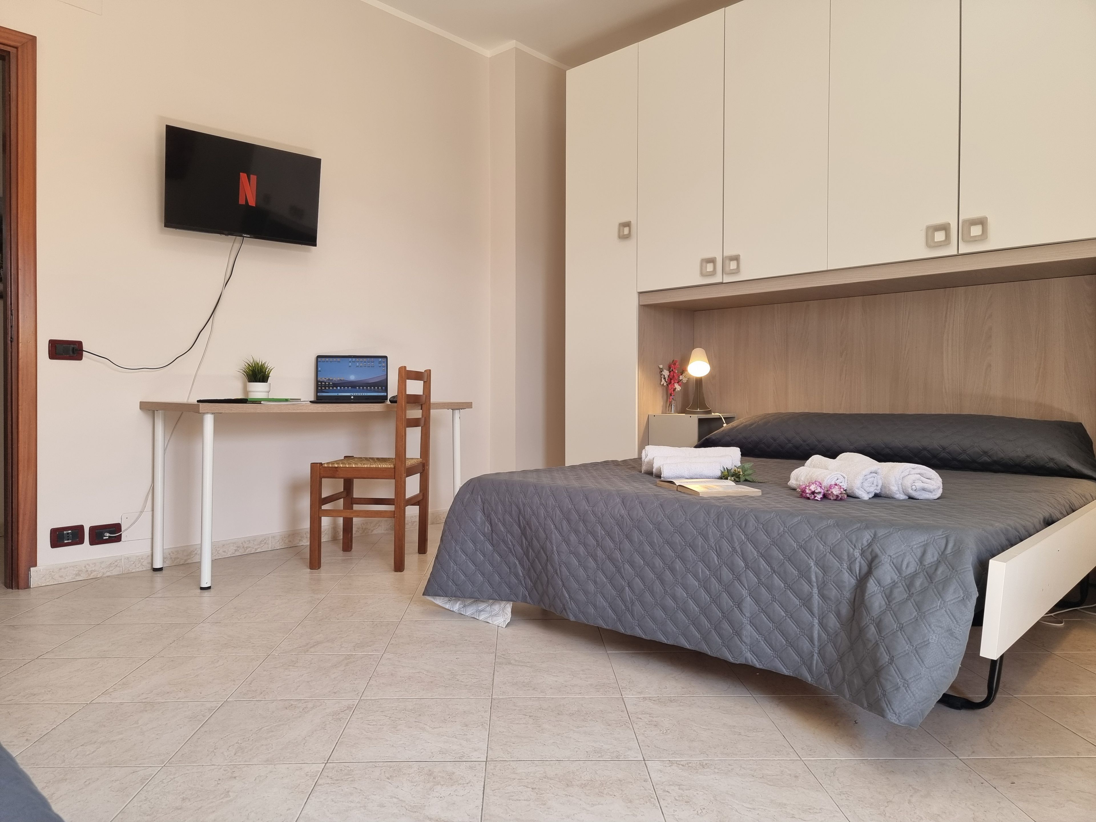 A bedroom with a bed, a desk with a laptop, a television mounted on the wall, and a nightstand with a lamp.