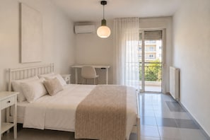 A hotel room with a bed, bedside tables, a desk, and a chair. There is a balcony with a view of buildings. - Luesh ΙΙ · Large 2BR Apt | Walk to Beach and Center (Agios Nikolaos)