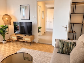 A living room with a flat-screen TV on a wooden stand, a sofa with a patterned cushion, a floor lamp, and a potted plant. - Stylish 1-Bedroom Apartment with a View of Zurich - Tilia 52 (Zürich)