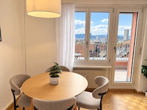 A round dining table with four chairs, a potted plant, and a window with a view of a cityscape. - Stylish 1-Bedroom Apartment with a View of Zurich - Tilia 52 (Zürich)