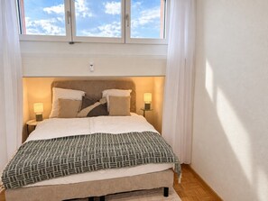 A bedroom with a bed, bedside lamps, a window, and a view of the sky. - Stylish 1-Bedroom Apartment with a View of Zurich - Tilia 52 (Zürich)