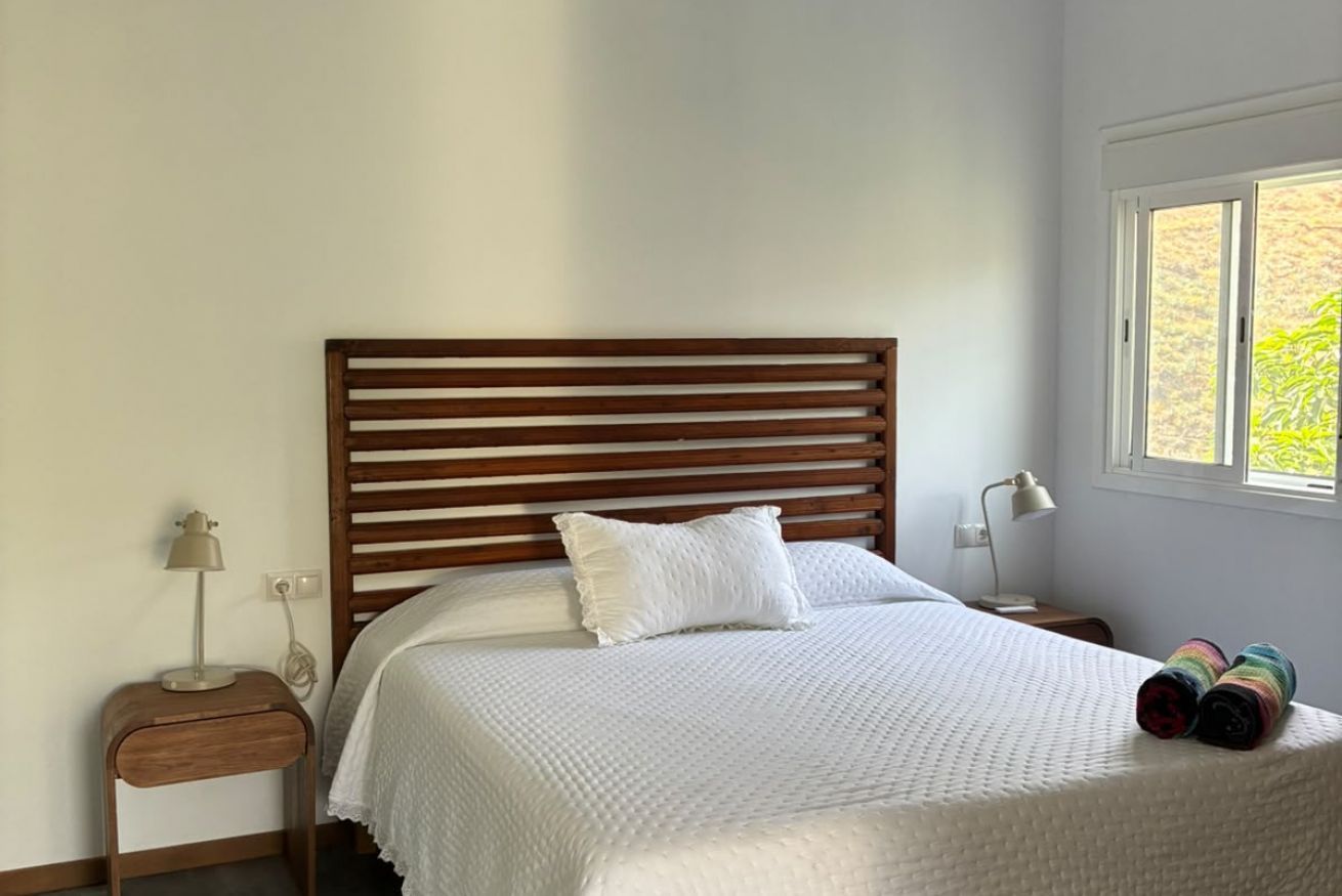 Une chambre à coucher avec une tête de lit en bois, une couvre-lit blanche et une fenêtre donnant sur un paysage verdoyant.