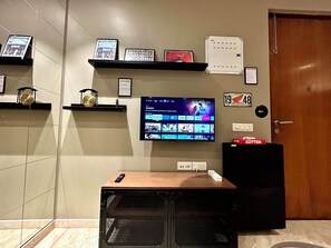 Een kamer met een wandgemonteerde flatscreen-tv, een plank met ingelijste foto's, een kleine speaker en een deur met een bordje.