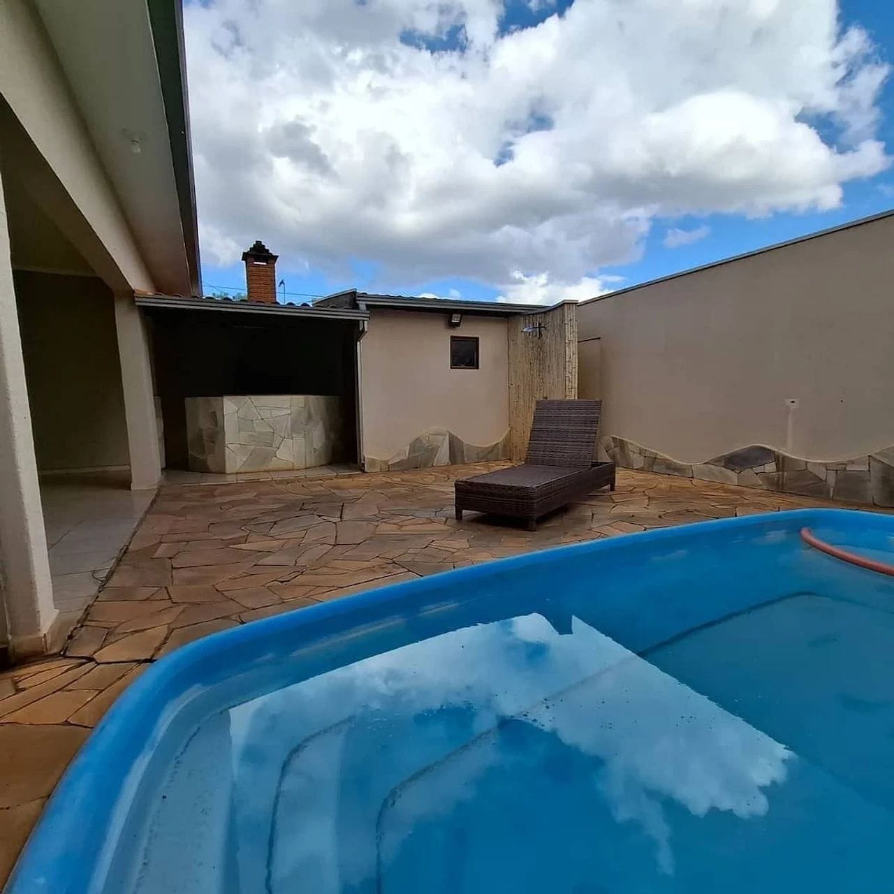 A swimming pool with a lounge chair, a stone patio, and a building with a chimney.