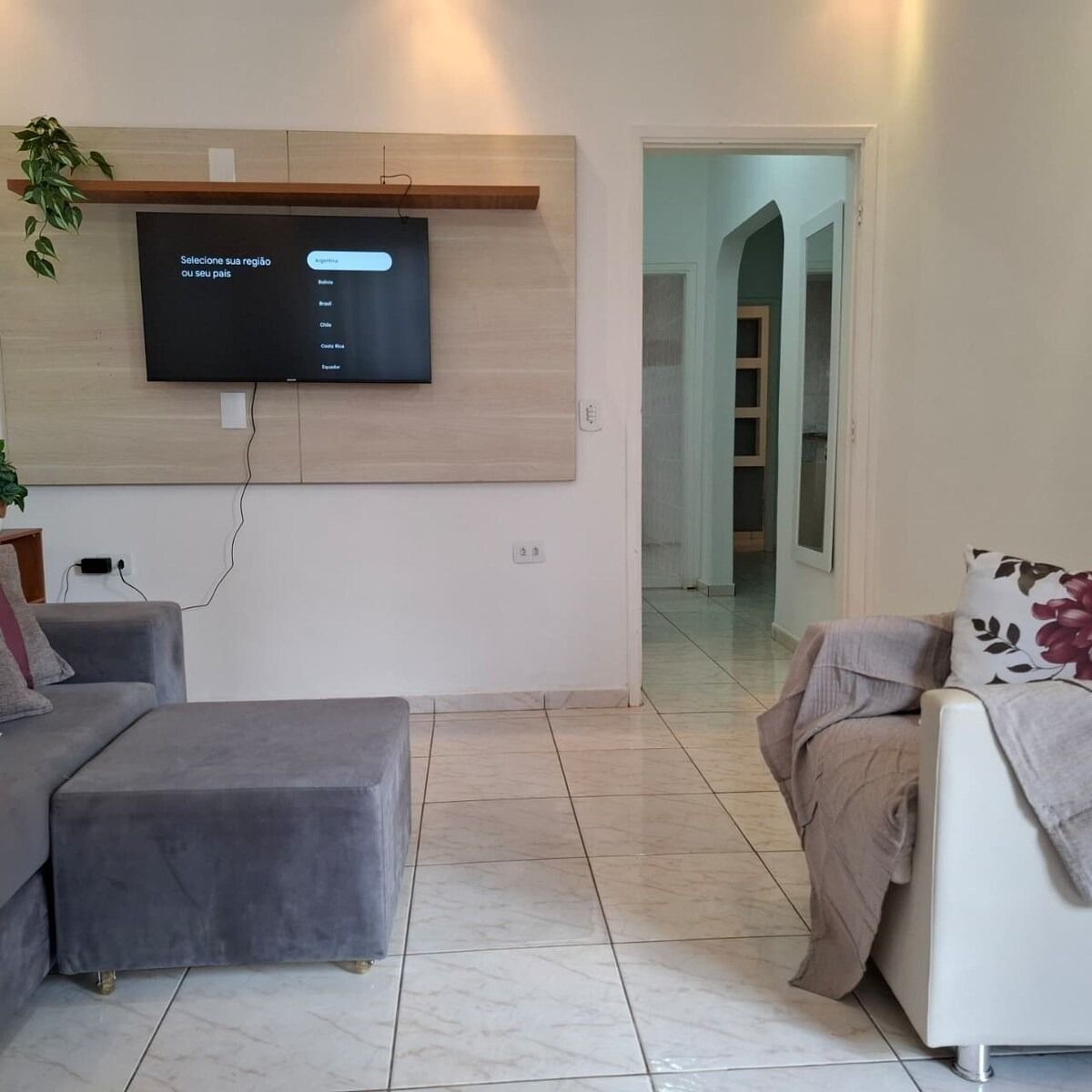 Uma sala de estar moderna com uma televisão de tela plana fixada em um painel de madeira, um sofá cinza, um sofá bege com almofada floral e um pequeno banquinho.