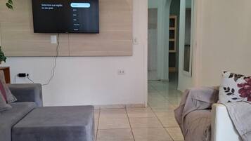 Uma sala de estar moderna com uma televisão de tela plana fixada em um painel de madeira, um sofá cinza, um sofá bege com almofada floral e um pequeno banquinho.