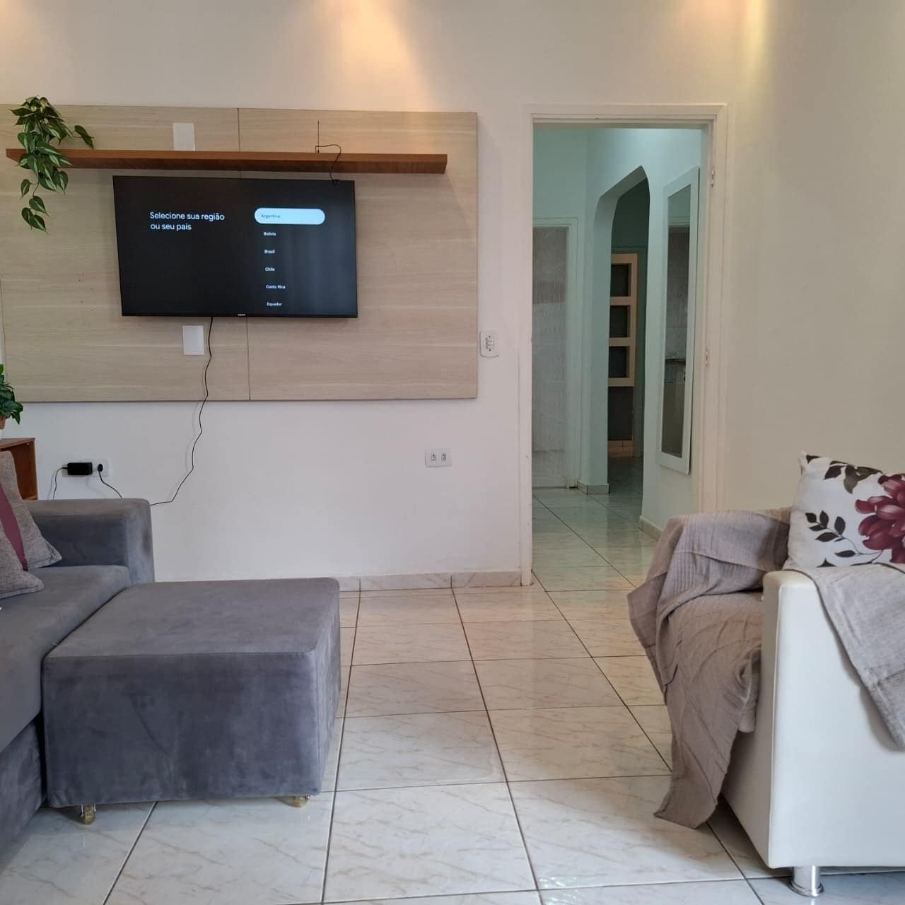 A modern living room with a flat-screen TV mounted on a wooden panel, a grey sofa, a white armchair, and a small table with a plant.
